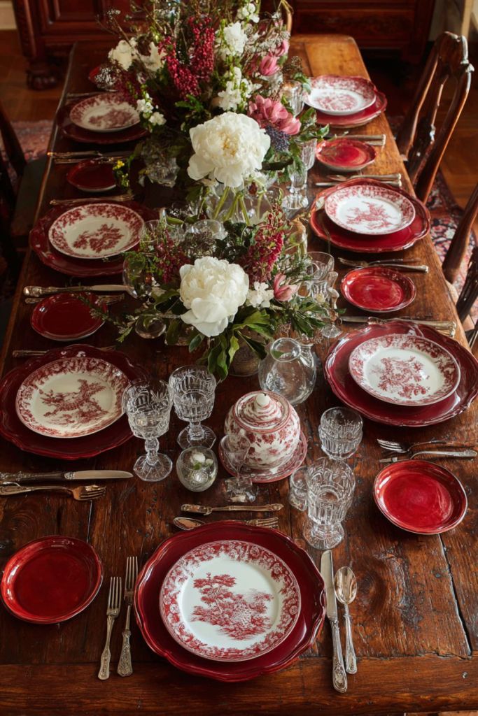 a rustic wooden dining table set with red and white floral china, crystal glasses, silver cutlery, and a floral centerpiece-thanksgiving table decor-loves from bet