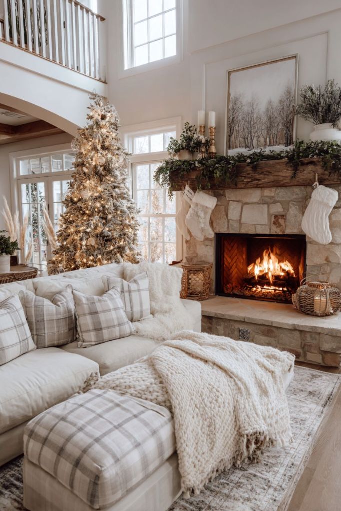 Neutral farmhouse Christmas living room with light stone fireplace, flocked tree, plaid pillows, knit throws, and greenery garlands for a cozy rustic look.