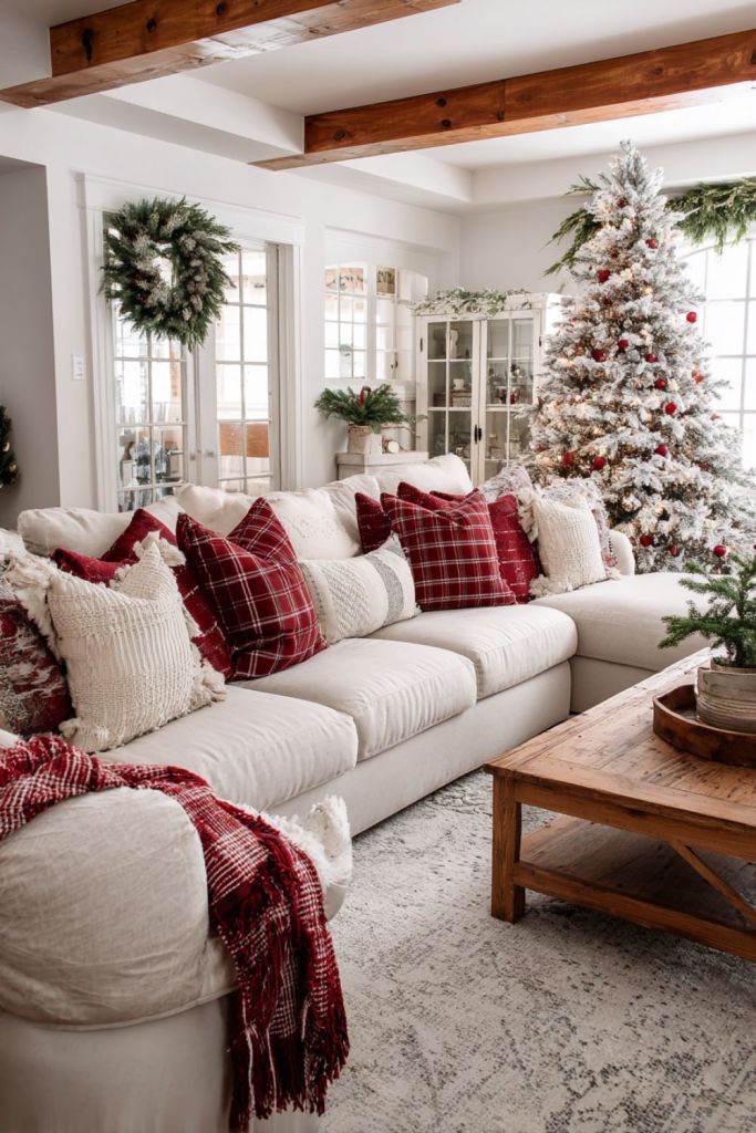 Cozy Christmas living room with white sectional, red plaid pillows, flocked tree, and wooden coffee table with greenery decor.